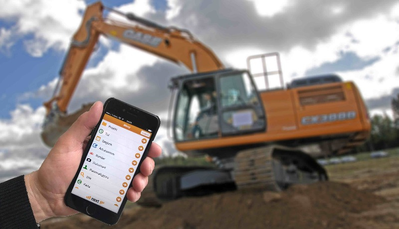 Hand som håller en mobiltelefon framför en grävskopa.